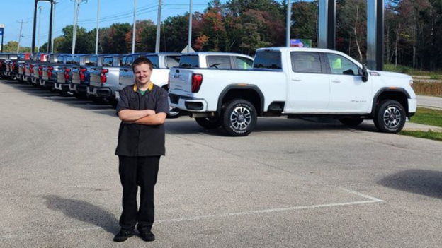 A confident young man stands in front of trucks at an auto dealer.